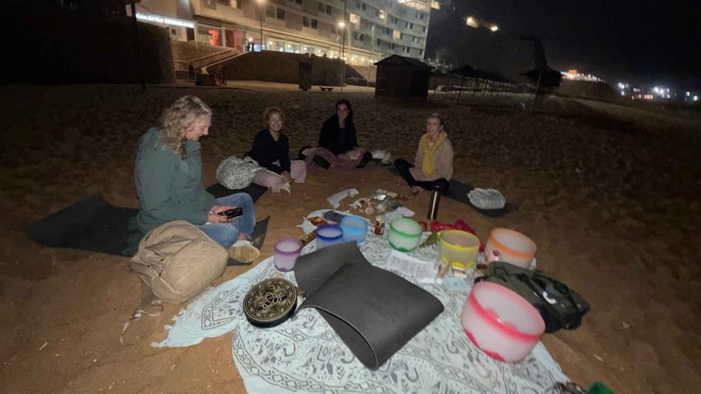 A group of people sitting on a blanket on a beach at night with various items scattered around. 1