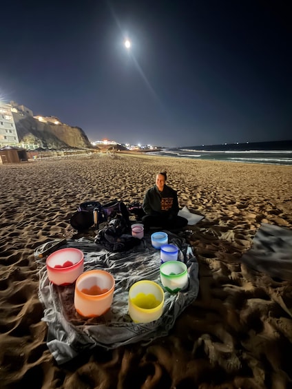 A person sitting on a blanket on a beach at night with a full moon in the sky and city lights in the background. 3