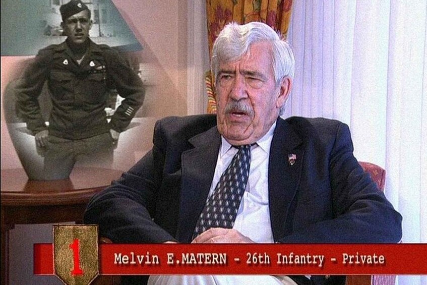 A man in a suit sitting in a room with a military uniform in the background. 3