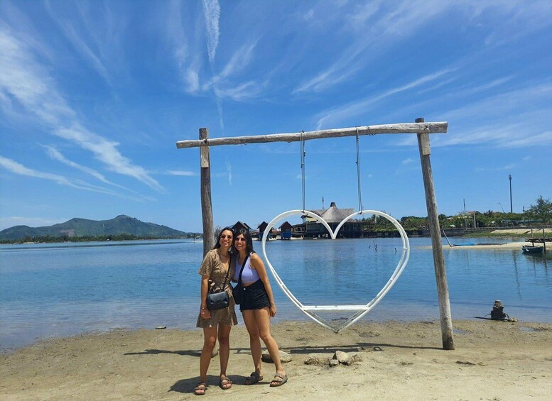 Two people standing in front of a heart-shaped swing structure by a body of water with mountains in the background. 5