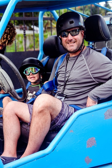 A man and a child in a blue vehicle with a steering wheel, both wearing helmets and safety harnesses. 3