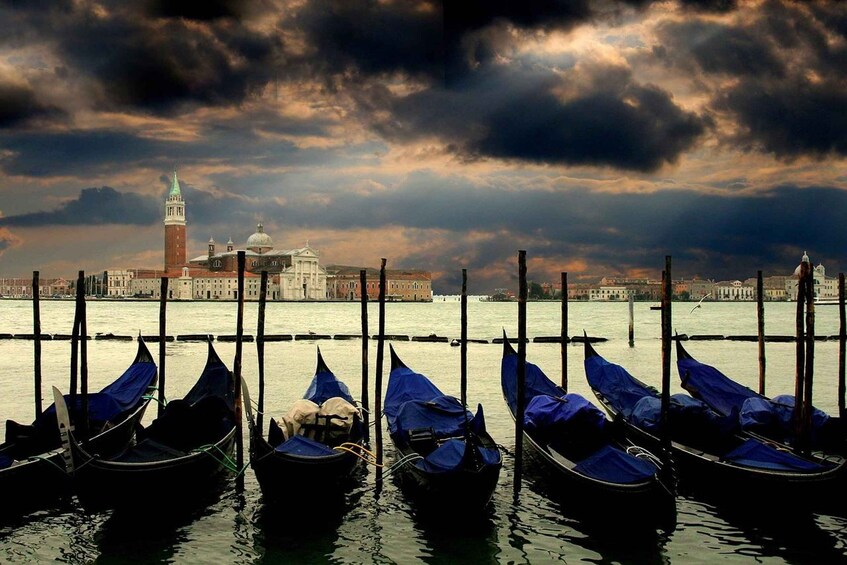 Gondolas docked at a pier with a historic building and a tower in the background under a cloudy sky. 3