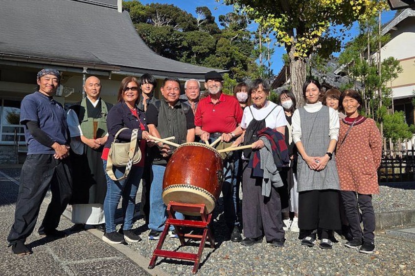 A group of people standing outdoors, some holding a traditional drum. 1