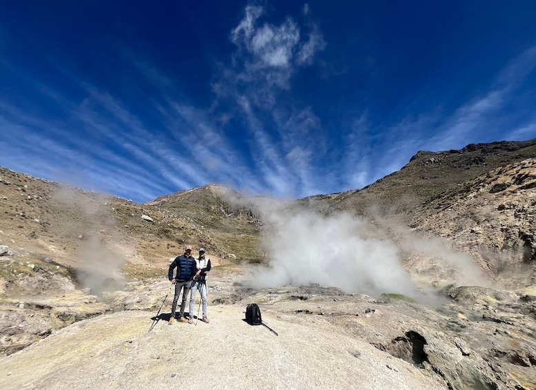 Two hikers on a rocky terrain with steam rising from the ground, surrounded by a mountainous landscape. 4