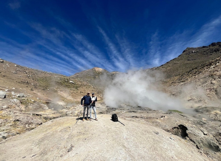 Two hikers on a rocky terrain with steam rising from the ground, under a clear blue sky. 3