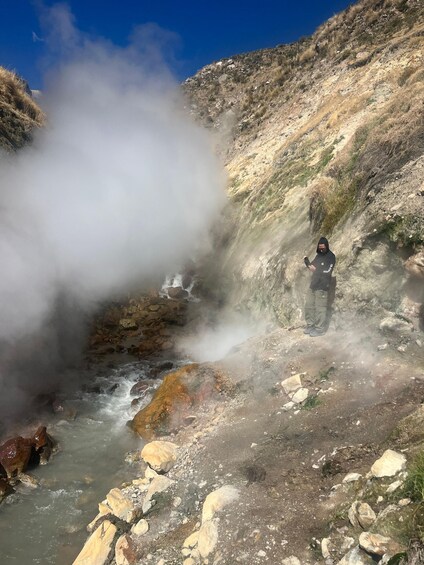 A person standing near a hot spring with steam rising from the water, surrounded by rocky terrain and sparse vegetation. 4