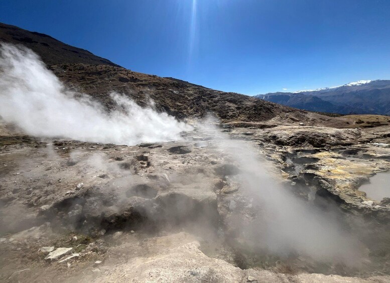 A geothermal area with steam rising from the ground, surrounded by rocky terrain and distant mountains. 1