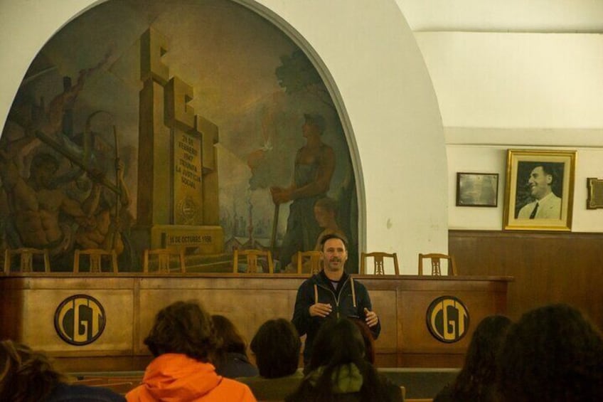 Un hombre hablando en un atril en una habitación con un mural y fotografías enmarcadas en la pared. 3