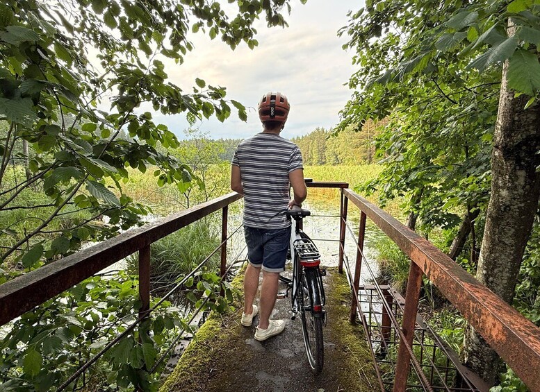 A person standing on a wooden bridge with a bicycle, surrounded by lush greenery and a river. 3