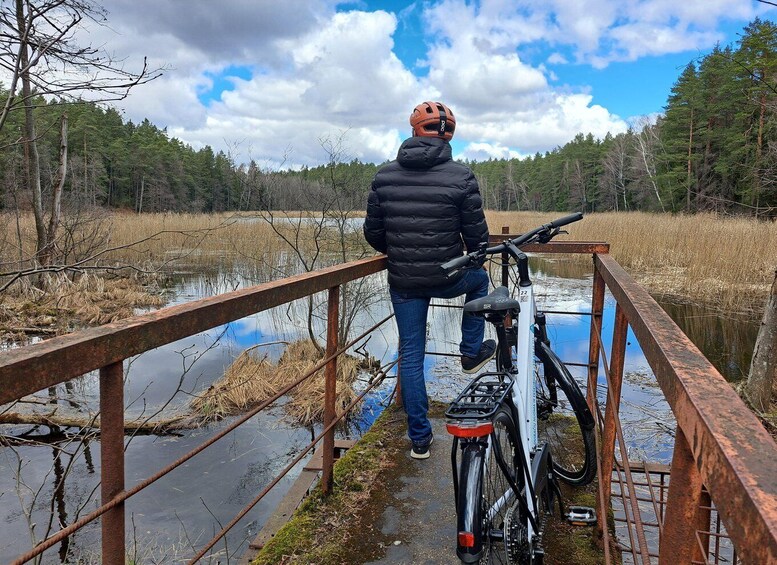 A person standing on a bridge with a bicycle, overlooking a body of water and forested area. 1