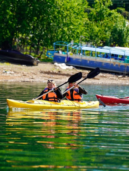 Two people kayaking on a calm river, surrounded by greenery and a dock with a boat. 2