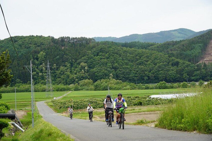 A group of cyclists on a paved path in a rural area with green fields and hills in the background. 5