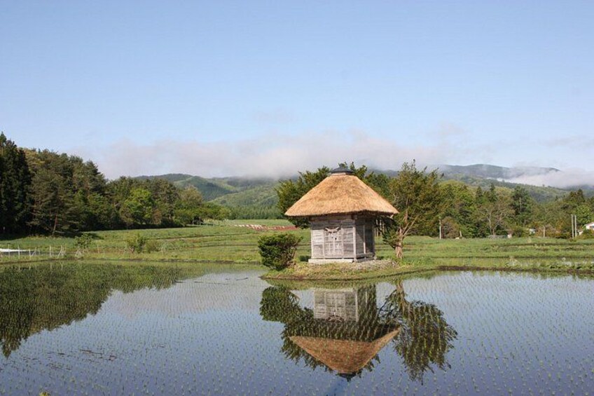 A thatched-roof hut by a calm pond with a reflection, surrounded by lush greenery and hills. 3
