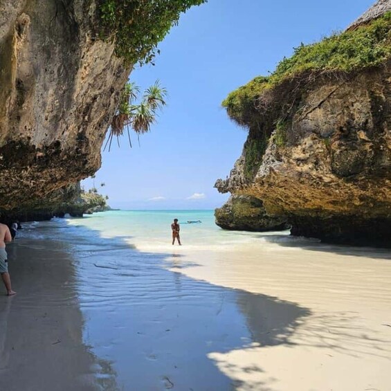 A beach with clear turquoise water, a person standing on the sand, and rocky cliffs with vegetation. 3