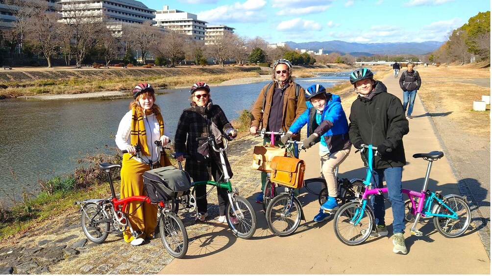 A group of people on bicycles by a river with buildings in the background. 1