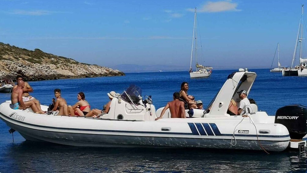 A small inflatable boat with people on it, anchored near a rocky coastline with sailboats in the background. 5