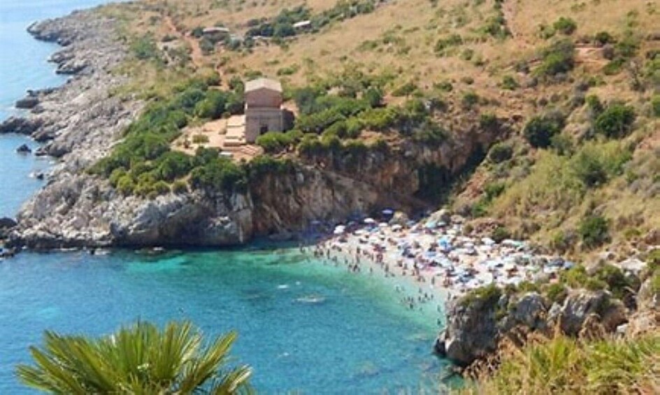 A coastal landscape with a rocky cliff, a small building, and a beach with people. 2