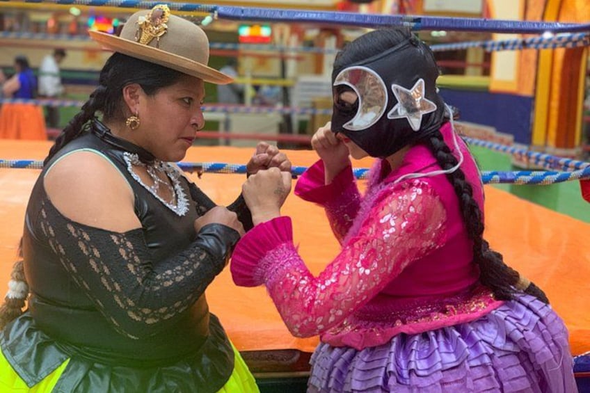 Two people in traditional attire, one in a black dress and the other in a pink top and purple skirt, engaged in a wrestling match. 1