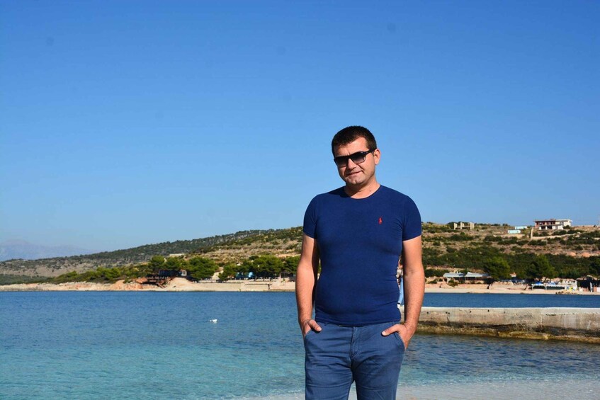A man standing by a clear blue sea with a hilly landscape in the background. 1