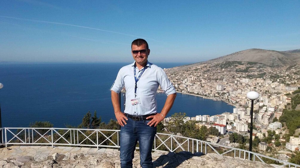 A man standing on a rocky ledge overlooking a coastal city with a clear view of the sea and a distant mountain. 5