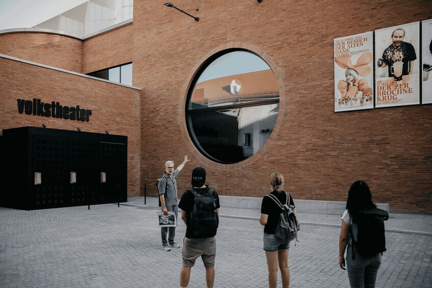 A brick building with a large circular window reflecting a building and a Volkstheater sign. 4