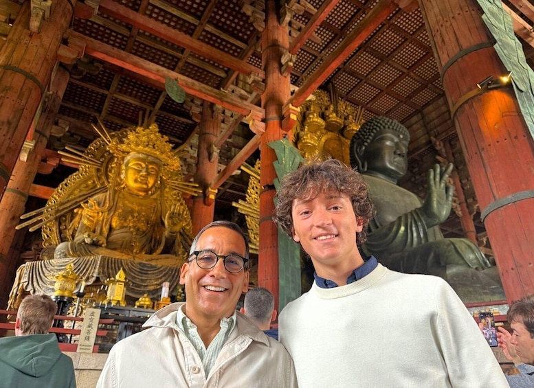 A man and a woman in front of a large golden Buddha statue in a temple with wooden architecture. 1