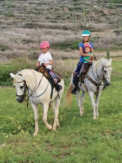 Two children riding horses in a grassy field with sparse vegetation and a cactus in the background. 4