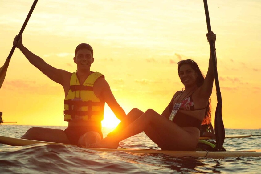 Two people sitting on a surfboard in the ocean at sunset. 2