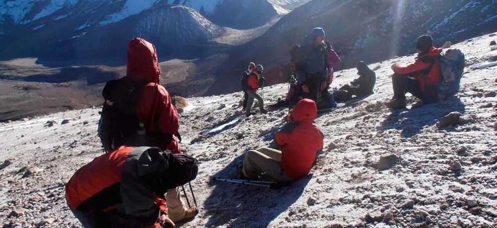A group of people in red jackets sitting and standing on a snowy mountain slope. 2
