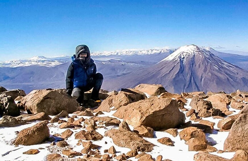 A person crouching on rocky terrain with a snow-capped mountain in the background. 5