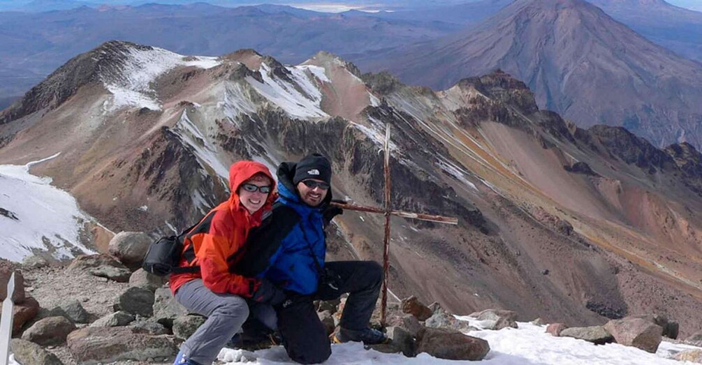 Two people sitting on a snowy mountain peak with a clear view of a range of mountains in the background. 1