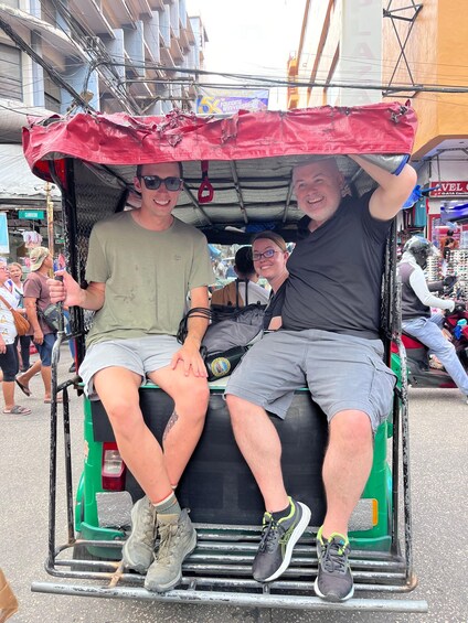 Two people sitting in a tuk-tuk with a red canopy, parked on a busy street with buildings and shops in the background. 1