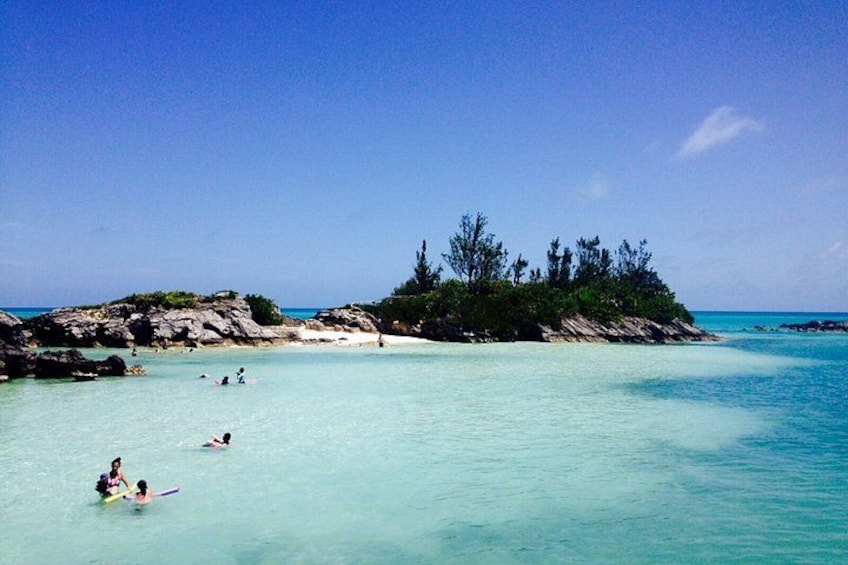 A beach with clear turquoise water, people swimming and surfing, and a small island with trees. 1