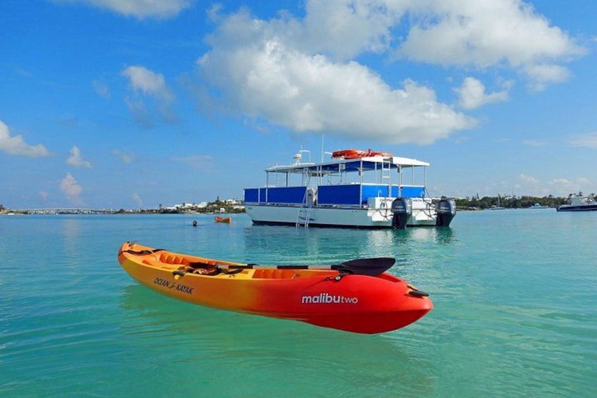 A red kayak with 'Ocean Kayak' and 'Malibu' written on it, floating in clear turquoise water. In the background, there is a large blue and white boat with an orange canopy. 2