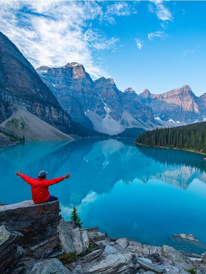 A person in a red jacket sitting on a rock by a calm lake with mountains in the background. 5