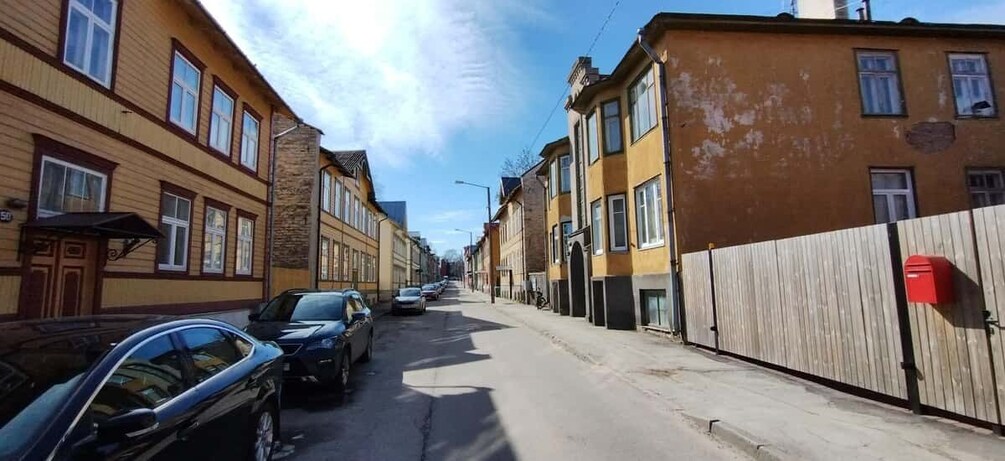 A narrow street lined with multi-story buildings, parked cars, and a wooden fence on one side. 3