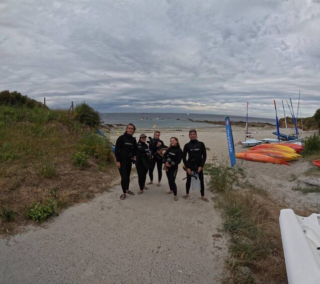 A group of people in wetsuits on a sandy path by the sea, with boats and a blue flag in the background. 5