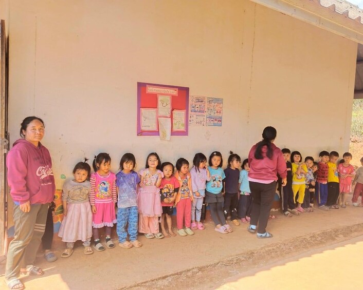 A group of children standing in a line on a concrete surface, with a wall featuring posters and a door in the background. 5