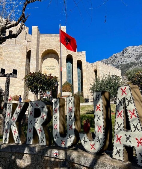 A building with a flag, the word 'KRUGA' in large letters, and a mountainous background. 1