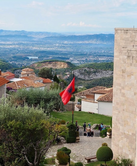 A red flag on a pole, a stone building, and a view of a valley with houses and hills. 3