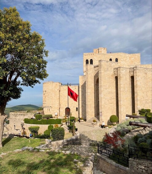 A historic stone building with a flag, surrounded by well-maintained gardens and a large tree. 5