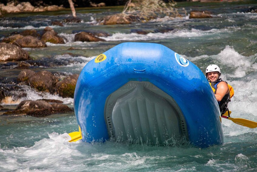 A person in a blue inflatable raft with a logo, paddling through a river with rocks and rapids. 4
