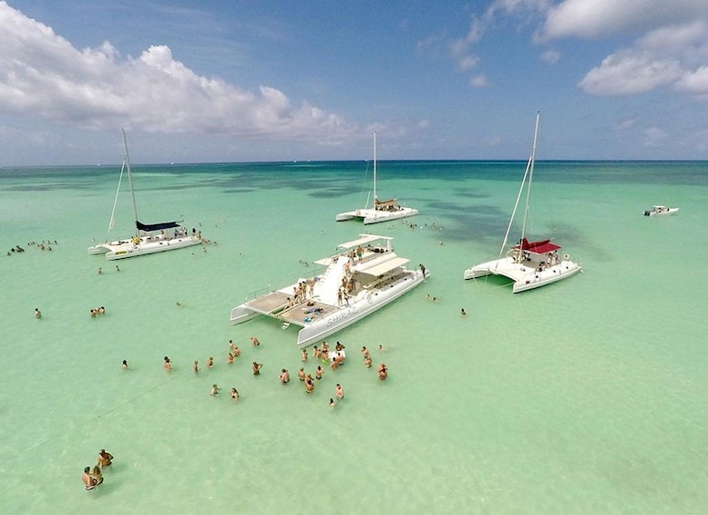 A tropical beach with several sailboats anchored in clear turquoise waters, surrounded by people swimming and sunbathing. 1