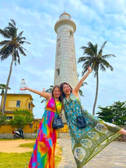 Two people posing in front of a lighthouse with palm trees and a yellow building in the background. 4