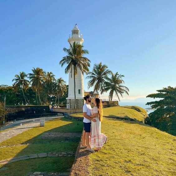 A couple embracing near a lighthouse with palm trees and a grassy hill in the background. 1