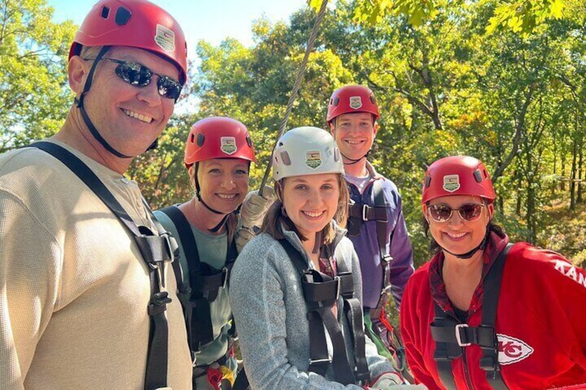 A group of people wearing helmets and harnesses, likely engaged in a zip-lining activity in a wooded area. 1
