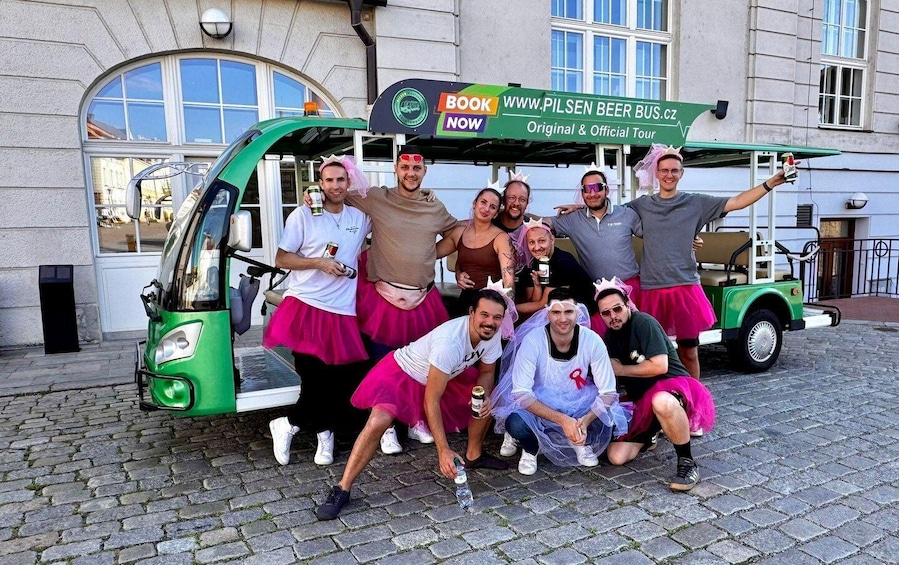 A group of people posing in front of a green vehicle with a sign that reads "BOOK NOW WWW.PILSENBEERBUS.CZ Original & Official Tour". 2