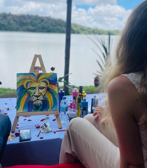 A person painting a colorful lion portrait outdoors by a lake. 1