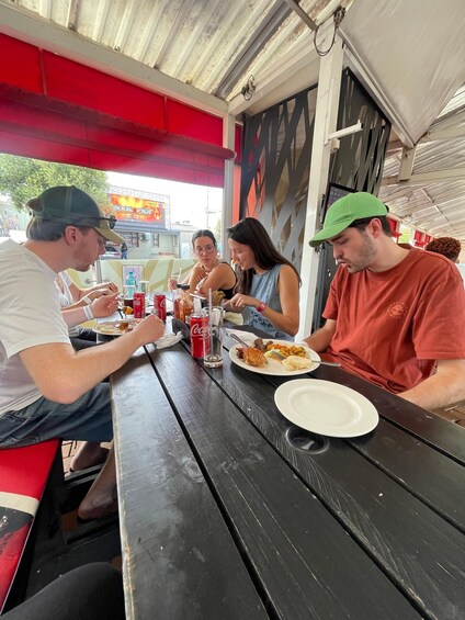Four people dining at an outdoor restaurant with a red awning and metal roof. 1