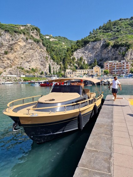 A boat docked at a pier with a man walking beside it, a rocky hillside, and buildings in the background. 2
